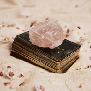 A stack of tarot cards with a rose quartz crystal on top, surrounded by petals on a fabric surface.
